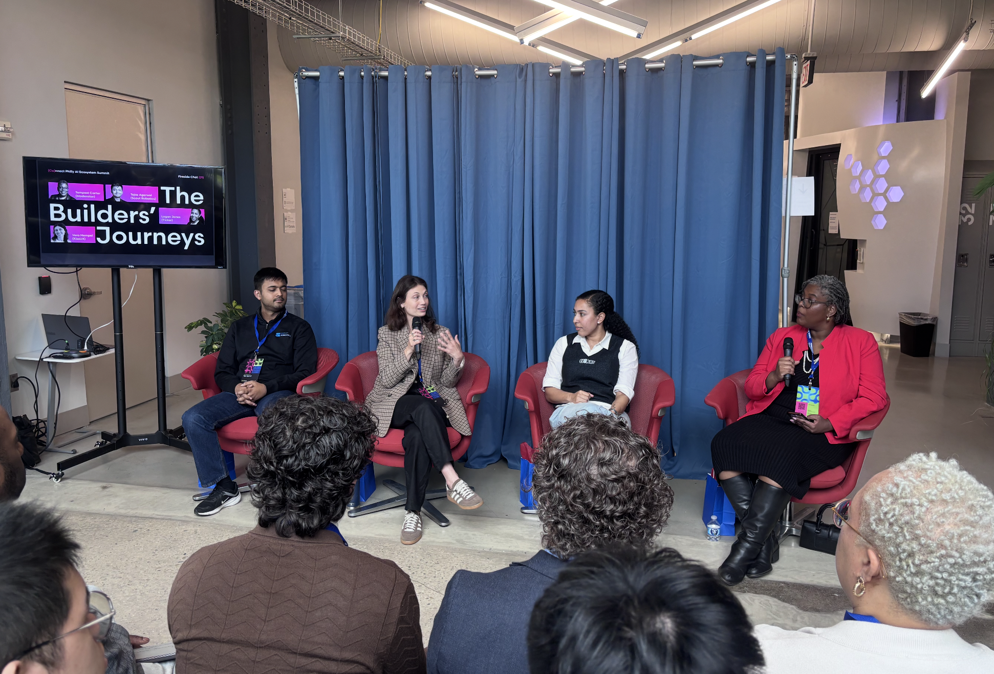 Vera speaking on a panel at the Philadelphia AI Ecosystem Summit, seated with three other panelists in red chairs in front of a blue curtain. A screen behind them reads 'The Builders' Journeys'.